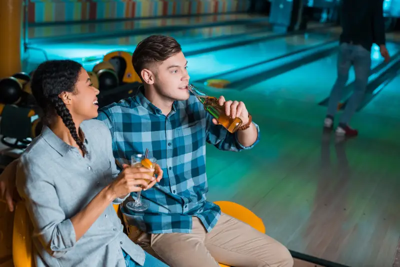 SPARE SOME ROOM FOR SNACKS AND DRINKS cheerful african american girl holding cocktail glass and talking to boyfriend drinking beer in bowling club